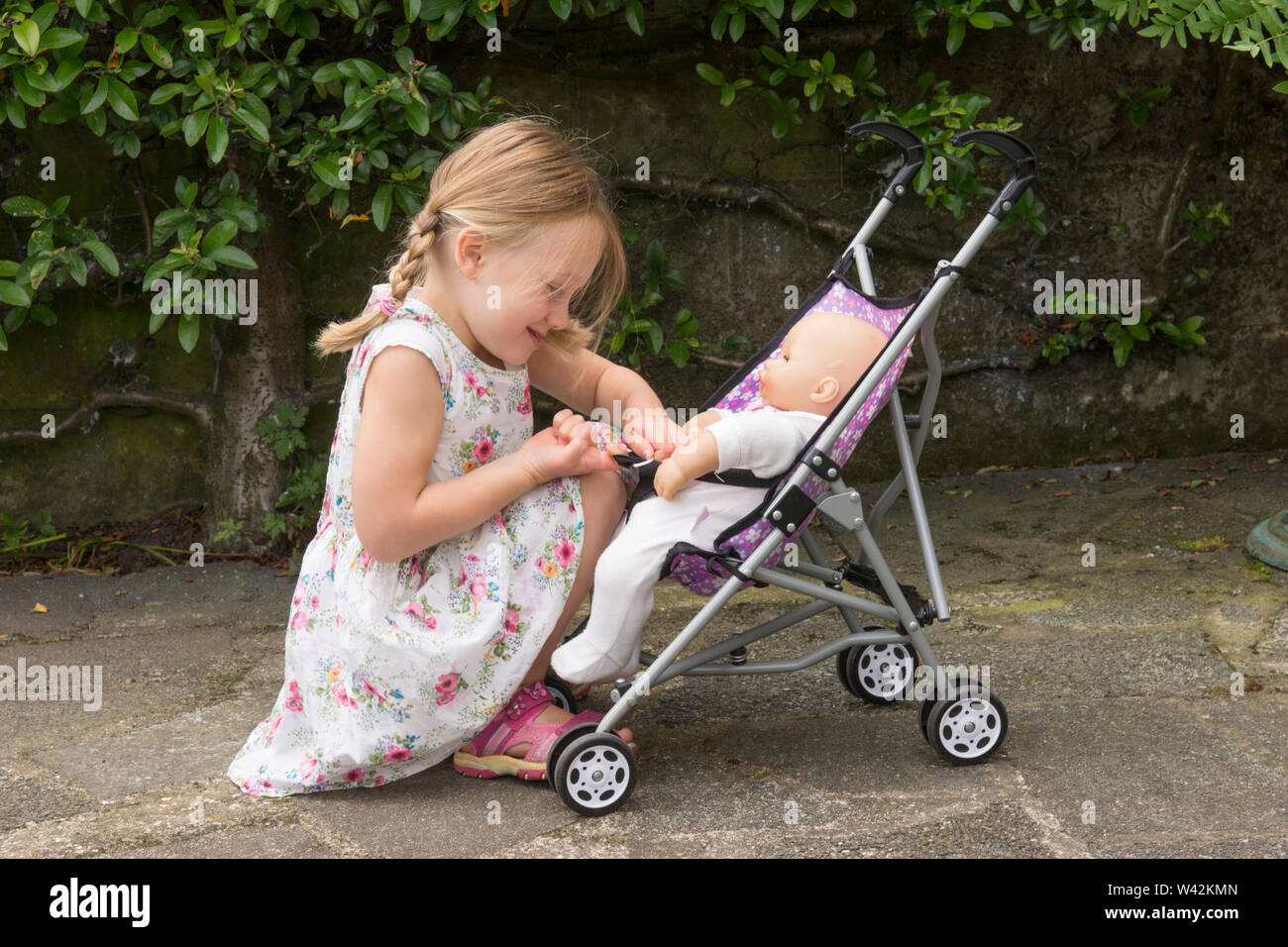 three year old child, young girl in pretty dress, fair hair in pig tails playing with doll in toy pushchair, outside in garden, UK Stock Photo