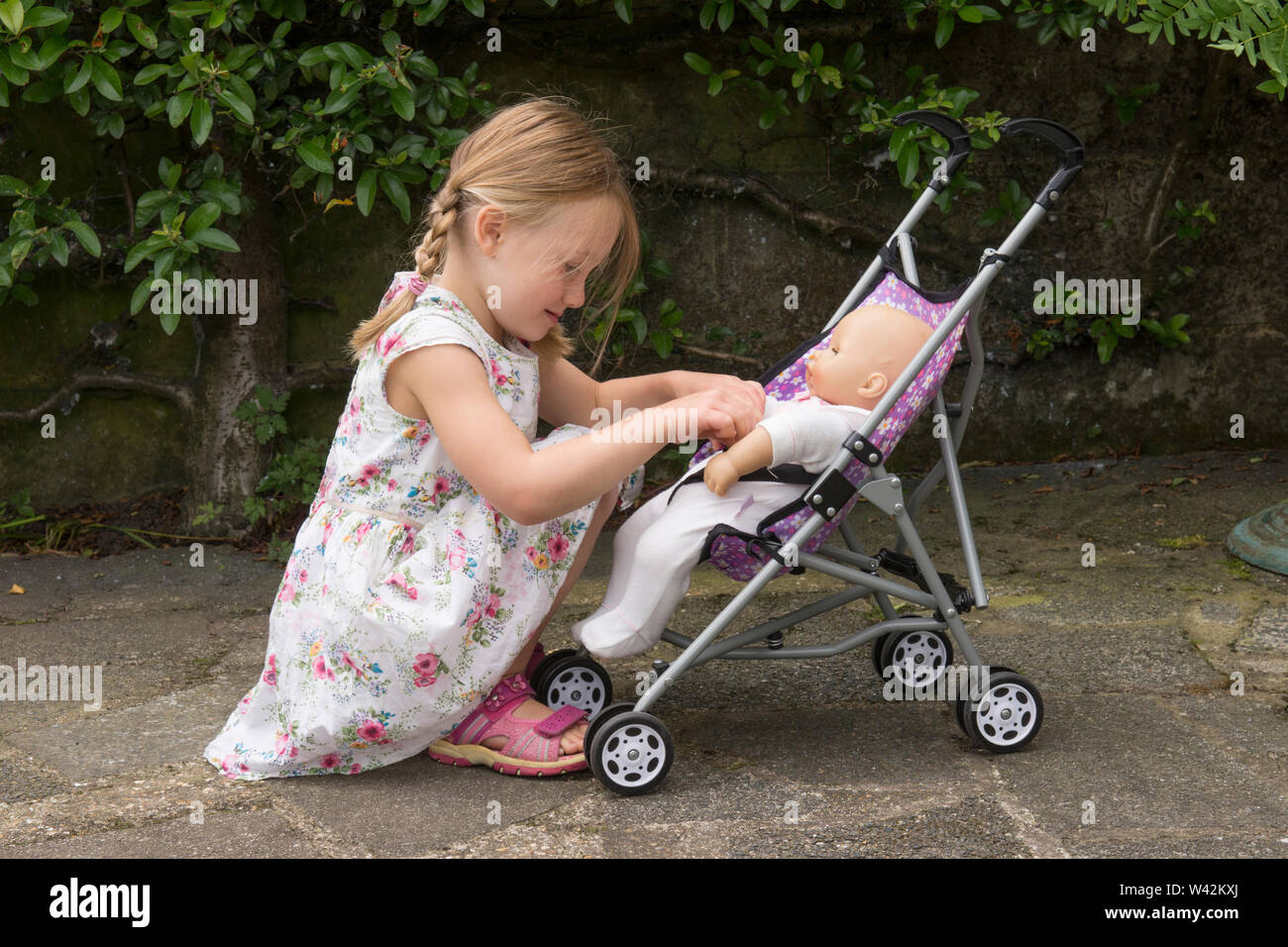 three year old child, young girl in pretty dress, fair hair in pig tails playing with doll in toy pushchair, outside in garden, UK Stock Photo