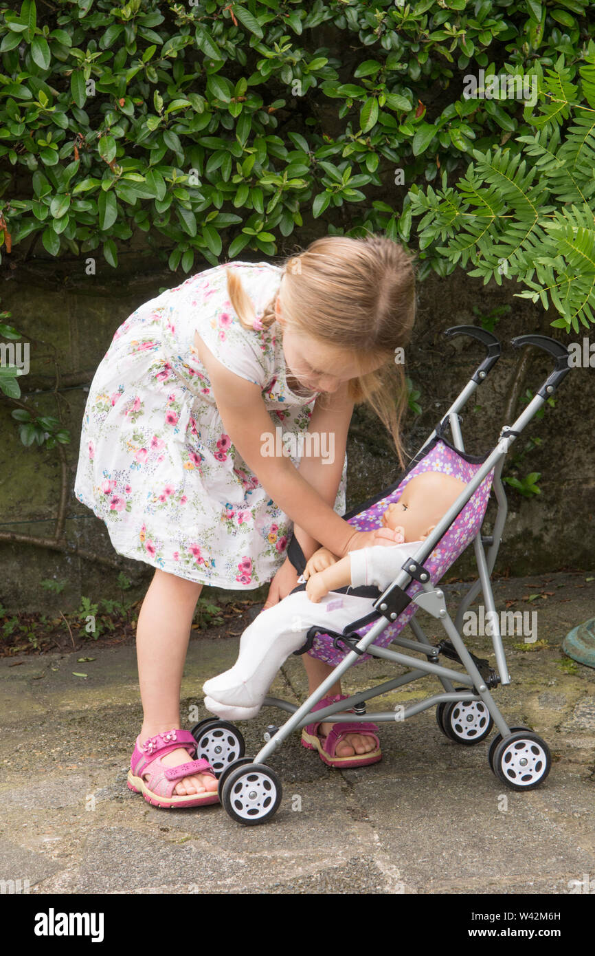 three year old child, young girl in pretty dress, fair hair in pig tails playing with doll in toy pushchair, outside in garden, UK Stock Photo