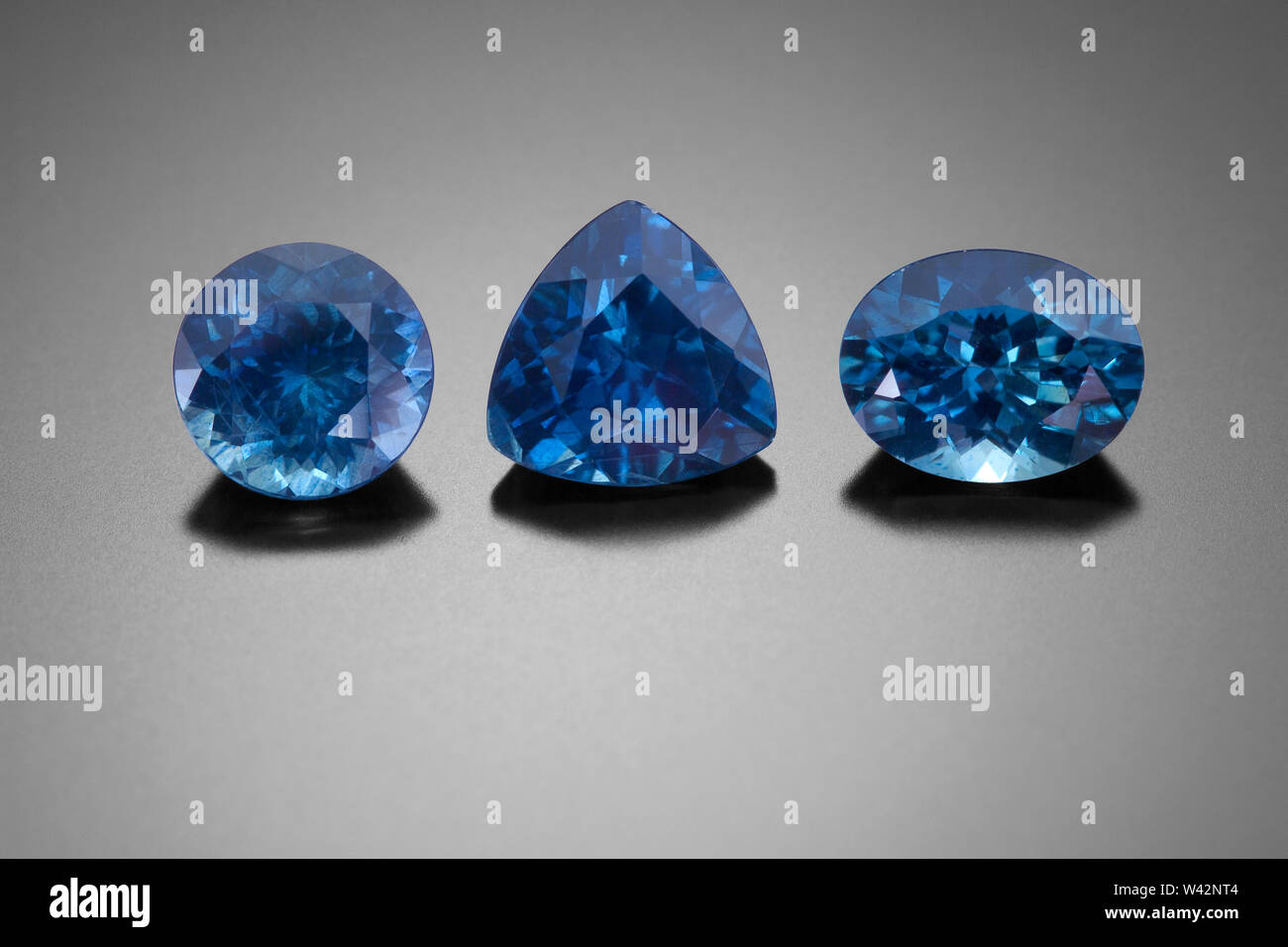 A faceted round, trillion and oval cut sapphires sit on a black reflective background. Stock Photo