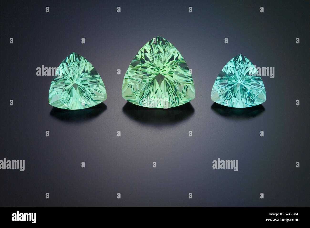 Three large fantasy cut pincushion triangle shape green tourmaline gemstone are shown on a dark reflective background. Stock Photo