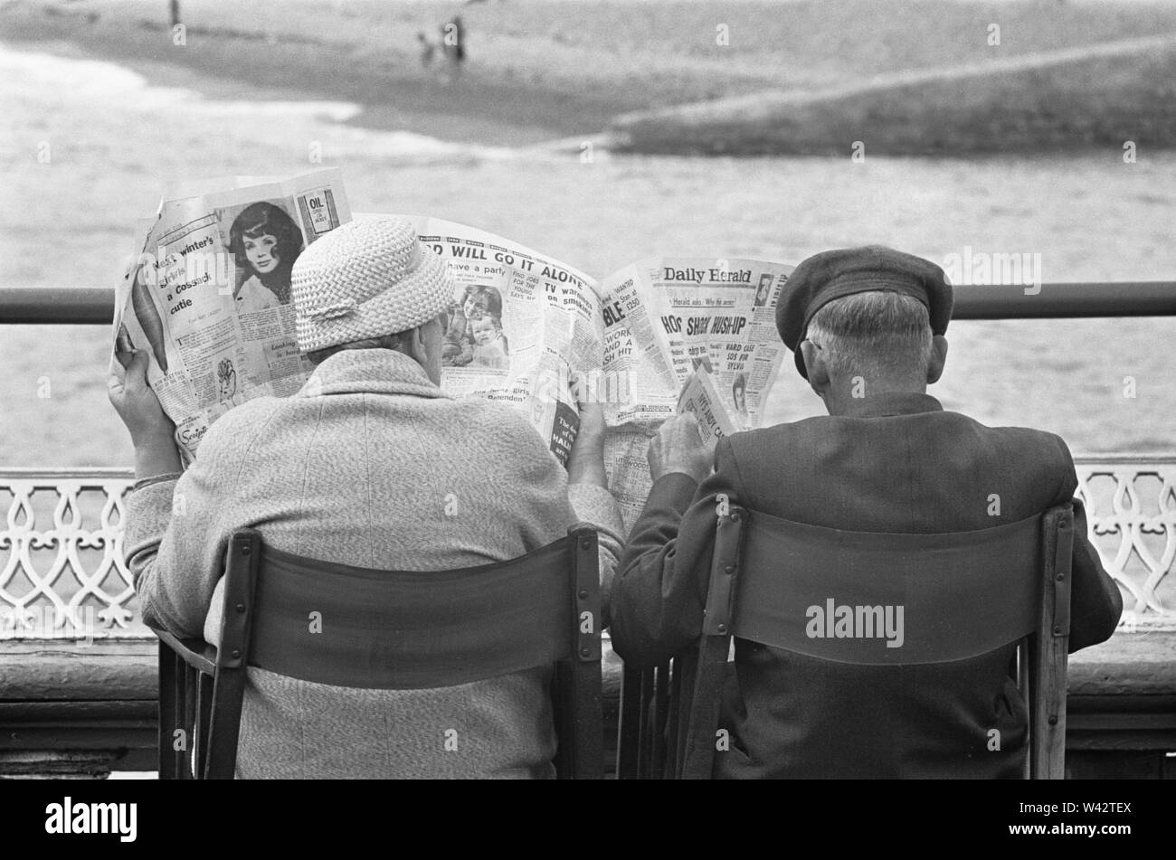 Holiday scenes at Brighton .Elderly couple reading the Daily Herald on Brighton Pier, whilst enjoying the sunny weather. 7th July 1963 Stock Photo