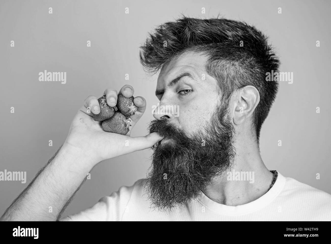 Hipster bearded holds strawberries fist as juice bottle. Man strict face enjoy fresh drink strawberry juice. Fresh juice concept. Man drinks strawberry juice suck thumb as drink straw blue background. Stock Photo