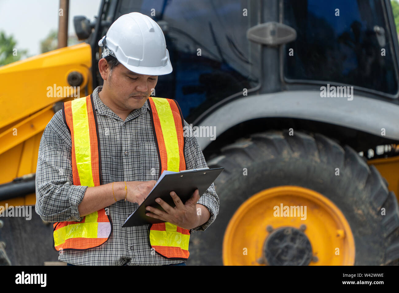 Engineering wearing a white safety helmet standing In front of the backhoe And are using tablet for check the blueprint with construction Stock Photo