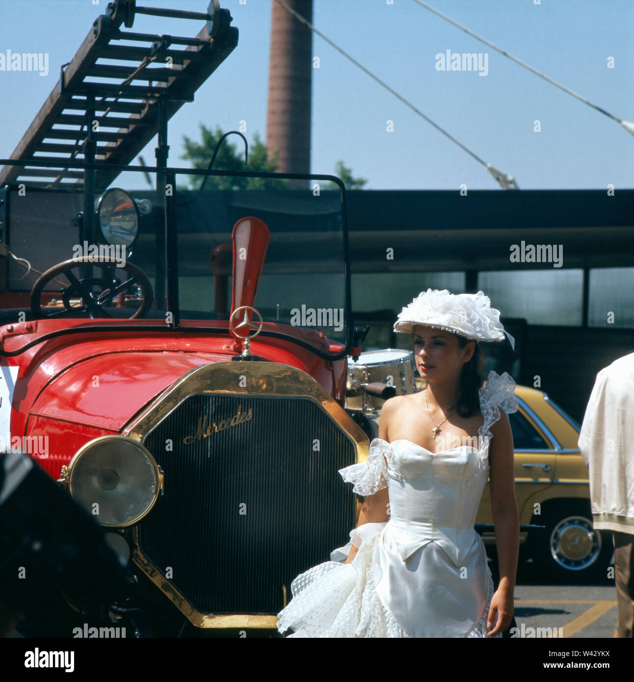 Eine junge Frau in einem Hochzeitskleid vor einem alten Mercedesmodell während einer Autoshow in München in den 1980er Jahren.  A young woman in a wedding dress in front of an old Mercedes model during a car show in Munich in the 1980s. Stock Photo