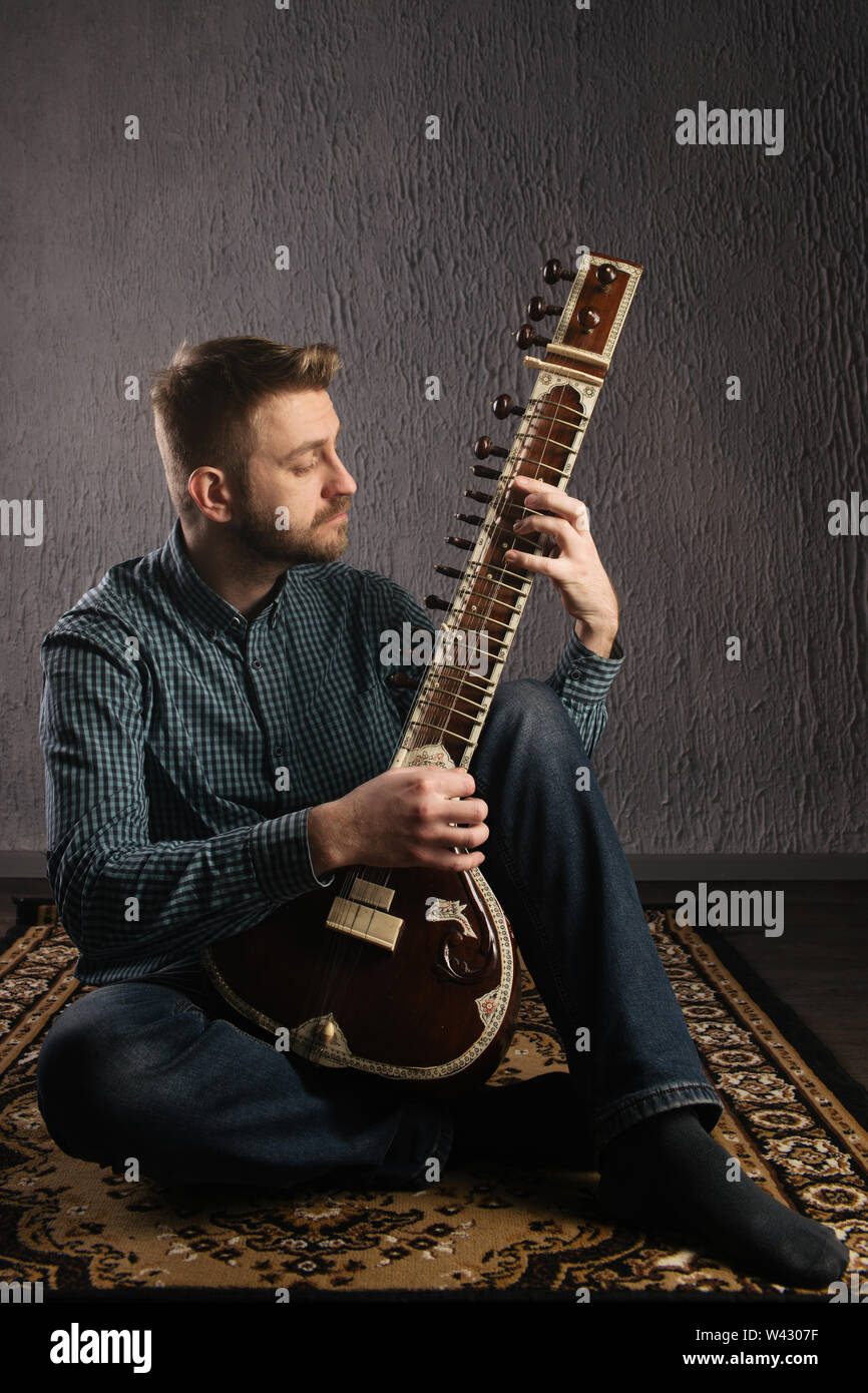 Portrait of a European man playing the sitar sitting on the carpet- Image Stock Photo