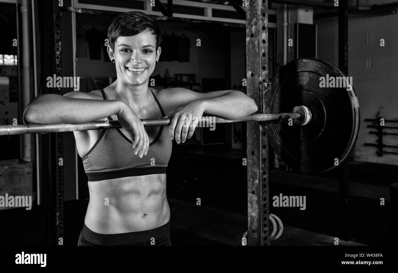 Portrait of an muscular beautiful woman with strong abs in a gym. The short haired female athlete is smiling and leaning against the barbell rack. Stock Photo