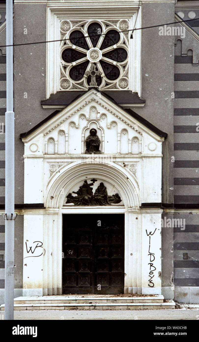 12th April 1993 During the Siege of Sarajevo: bullet and shrapnel holes and graffiti surround the entrance doors of the Church of the Holy Trinity on Sniper Alley (Zmaja od Bosne), in the Dolac Malta area of the city. Stock Photo