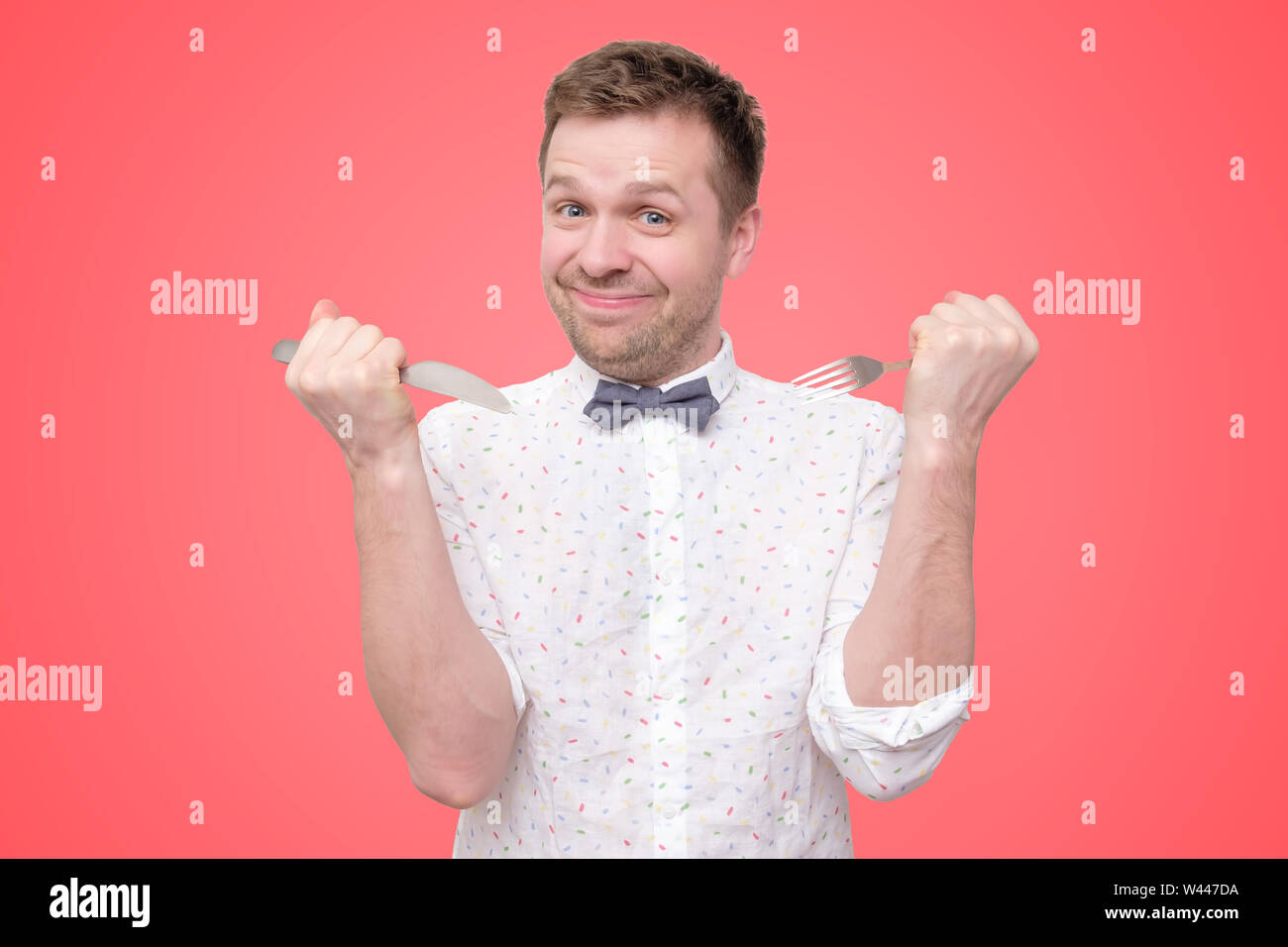 Hungry young man holding fork and knife on hand ready to eat, licking lips. Stock Photo