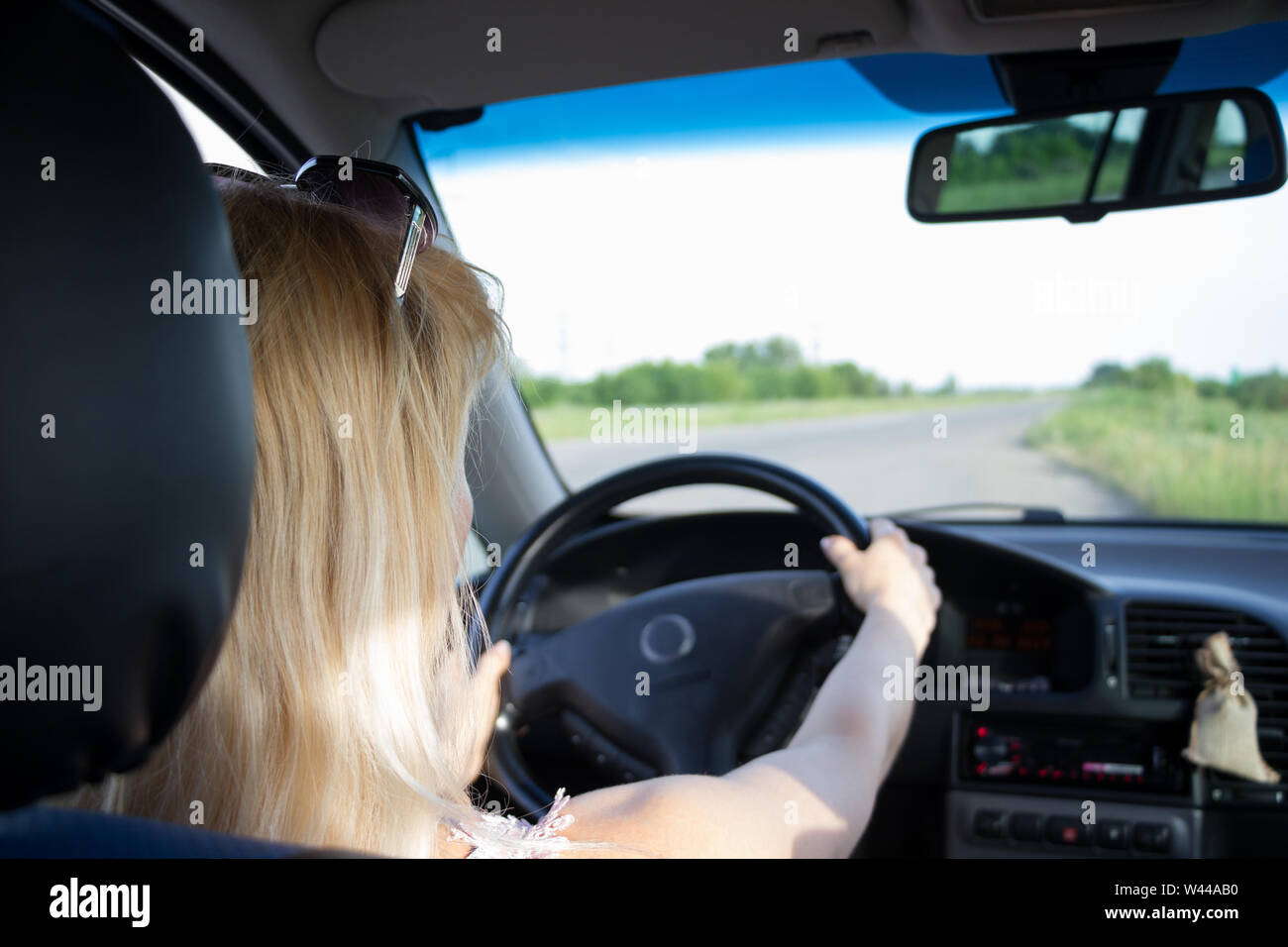 Attractive fair-haired girl keep both hands on steering wheel while driving an old car with black interior through the country road. Vehicle dashboard Stock Photo