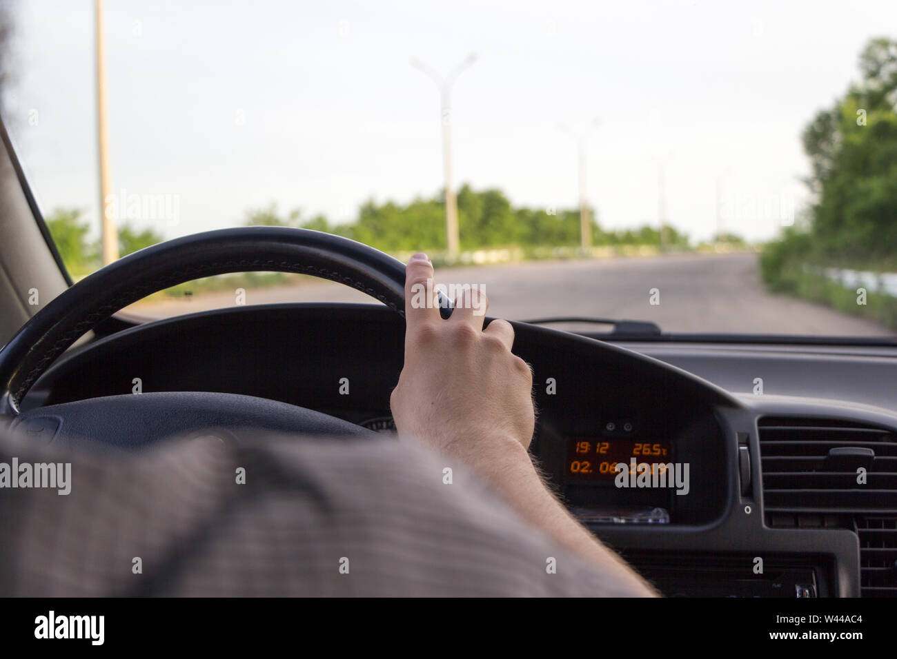 Mens hand keep hold on steering wheel while driving a car on road. View from behind. Safe driving concept. Summer day, green trees outside.  Black old Stock Photo