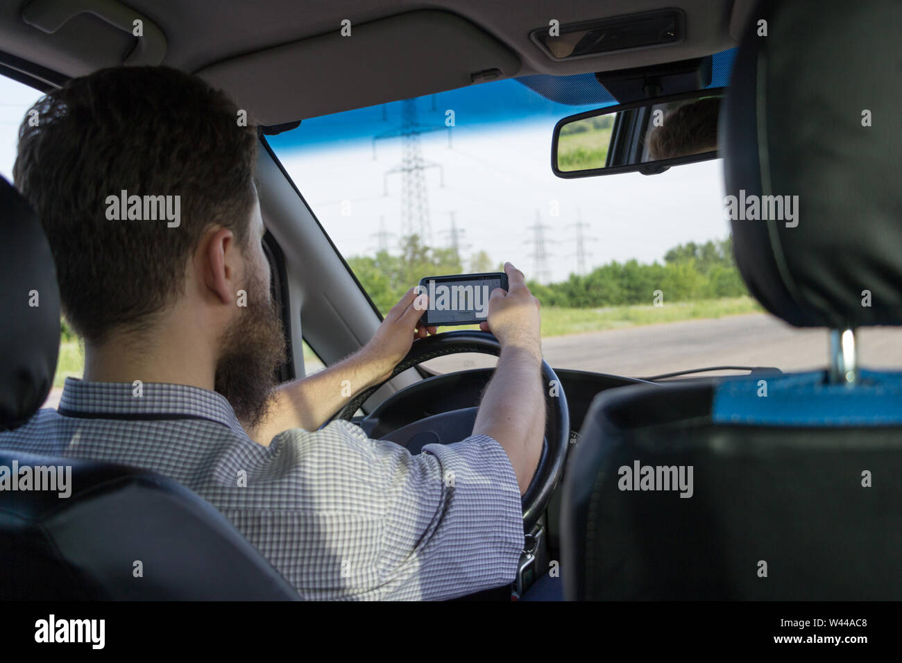 Young bearded man sitting behind the wheel and looking for route by navigation map in smartphone. Rear view. Safe journey concept. Driving car. Countr Stock Photo