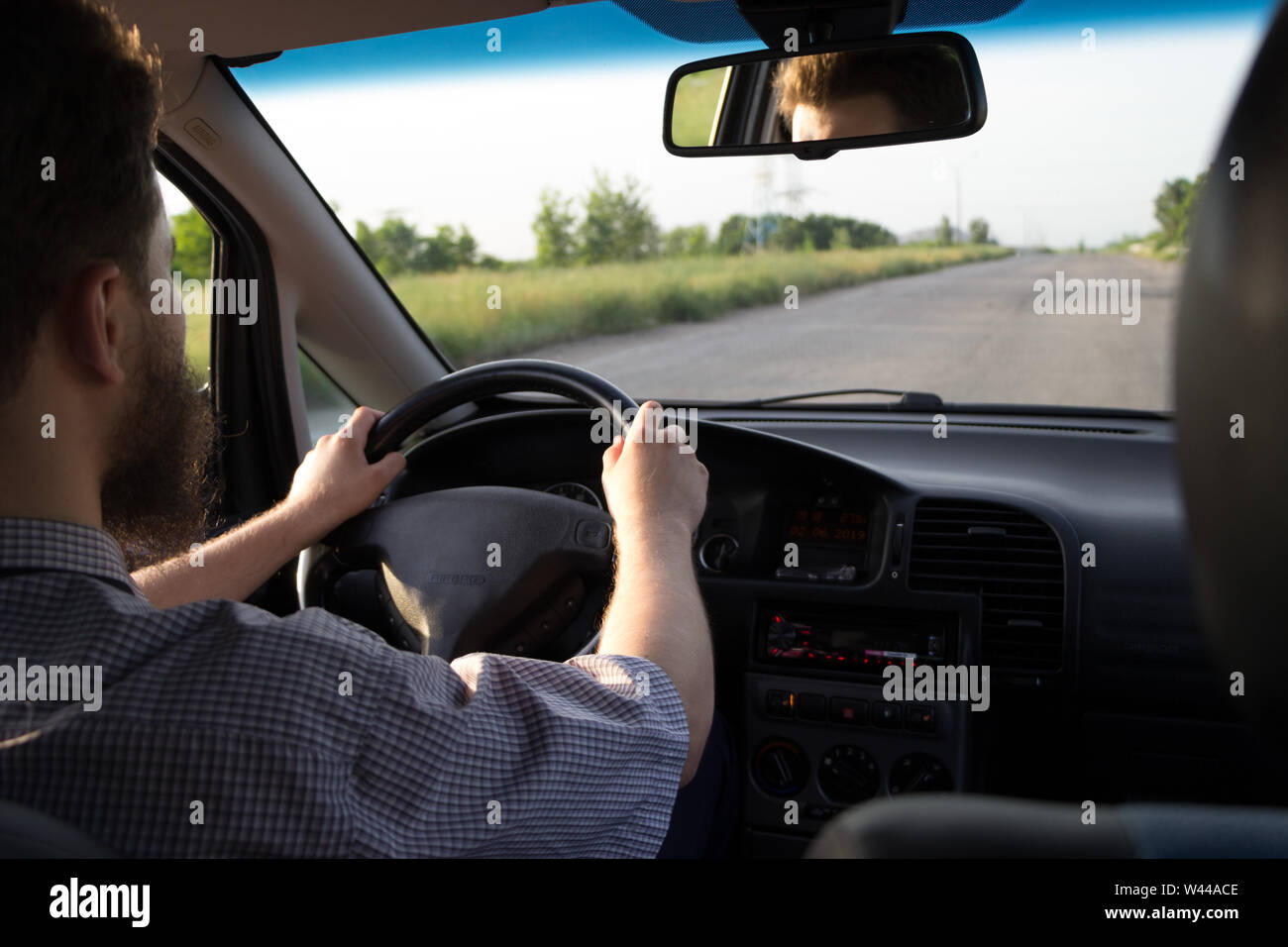 Young bearded man driving a car on old highway. Both hands holding a steering wheel. Safe driving concept. View from behind. Summer day, green trees a Stock Photo