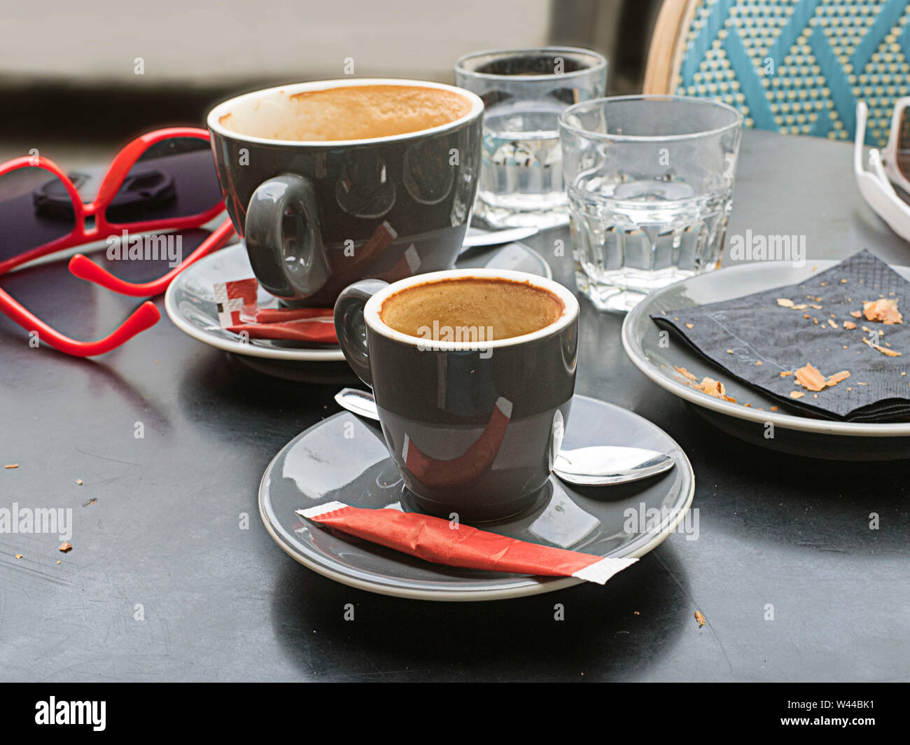 An outdoor cafe table after a couple's coffee break, showing two empty cups, a plate with the crumbs of a food item, and two pair of sunglasses, selec Stock Photo
