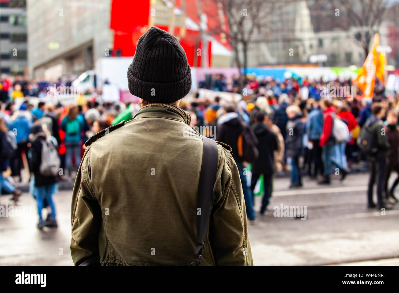A young man is seen from the back, wearing an olive jacket and wooly hat, as eco-activists march against global warming in a blurry background. Stock Photo