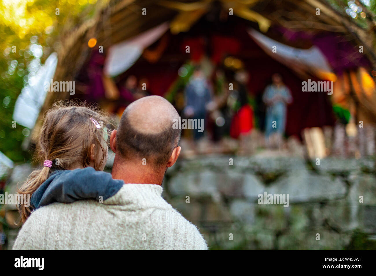 A bald headed father is viewed from behind, holding his his little girl as they watch a live band perform at a family music festival, with copy space. Stock Photo