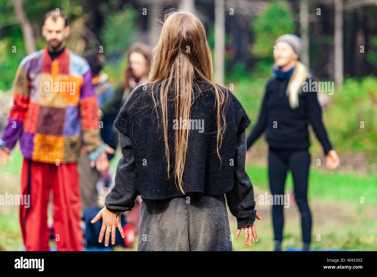 A girl with long blonde hair is seen from the rear, wearing native clothes and standing among a group of people performing spiritual meditation outdoors. Stock Photo