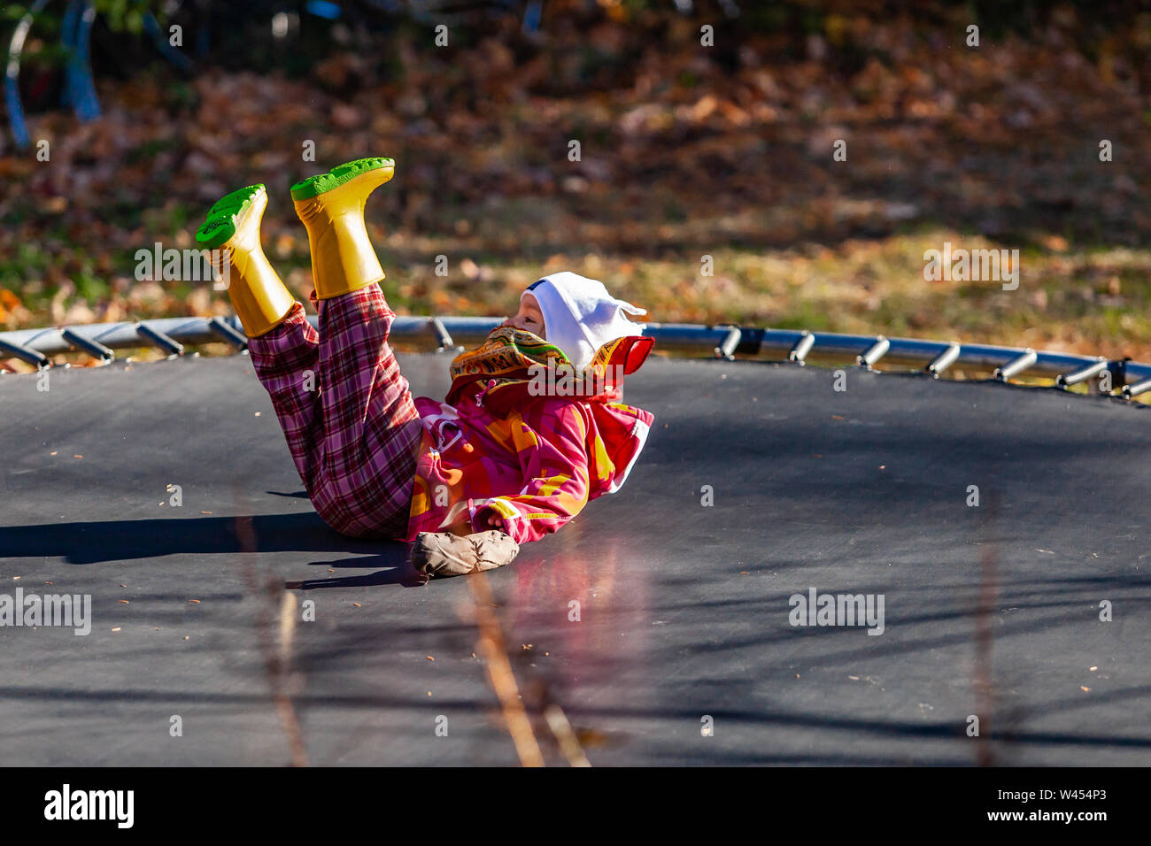 A closeup view of a young child, landing on her back whilst trampolining outdoors during autumn, wrapped up warm with yellow boots and pink jacket. Stock Photo