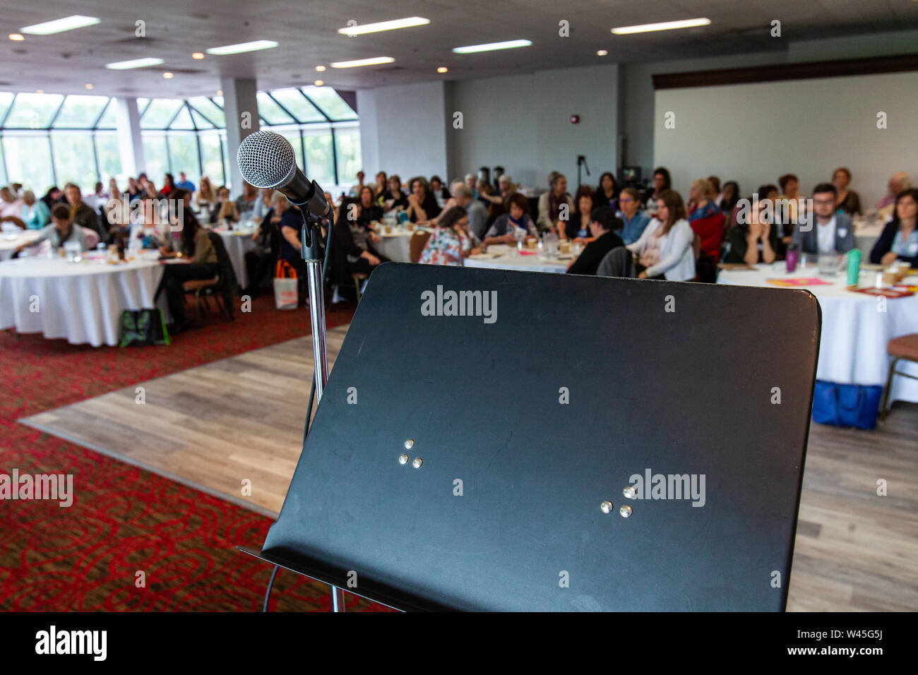 A speaker's stand with microphone is viewed close up during a packed conference for professional people, blurry employees are seen sitting behind. Stock Photo