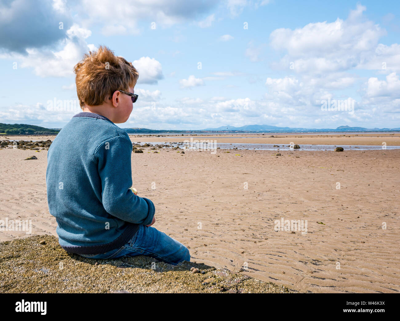 8 year old boy sitting on beach looking out to sea at low tide Firth of Forth, East Lothian, Scotland, UK Stock Photo