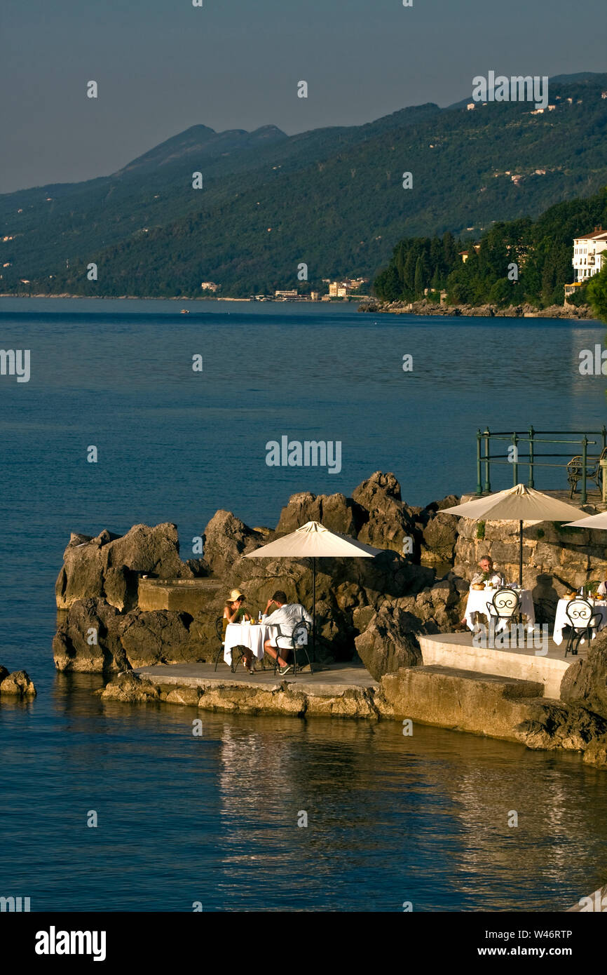 waterfront scene; people eating, tables, umbrellas, rocky prominence, peaceful, early morning, coastal, Lungomare; promenade; Adriatic Sea; Opatija; C Stock Photo