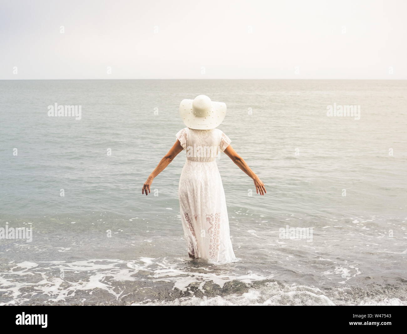 Adult woman wearing a white hat and dress walks on the shore of a beach ...