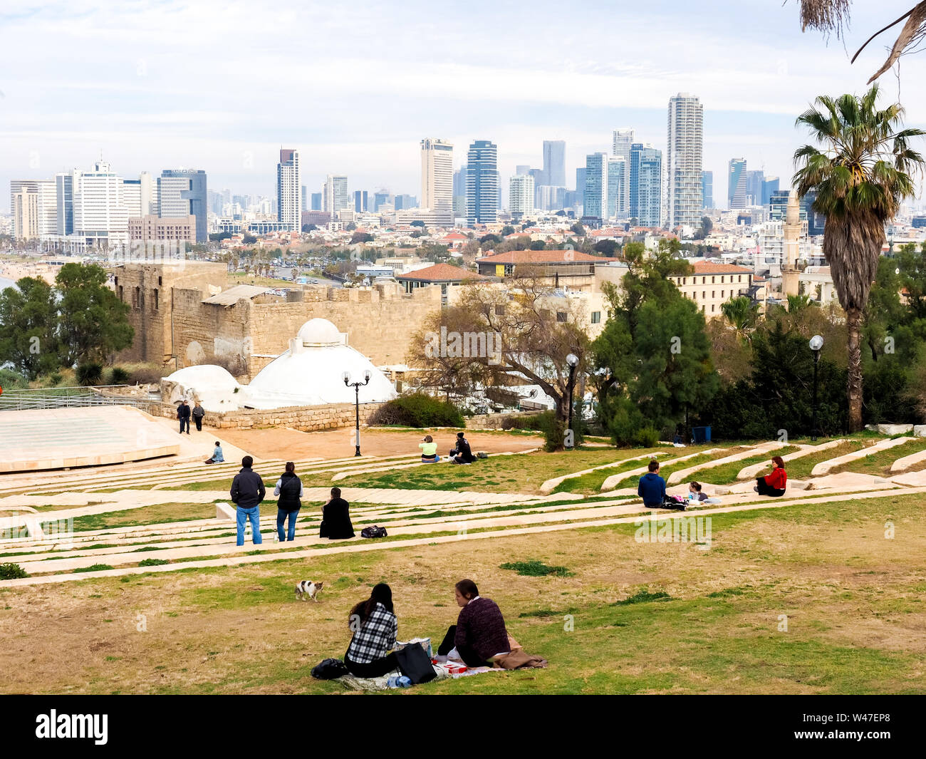 Jaffa, Israel - February 4, 2017: People relaxing on the lawn and admiring the view of the Tel Aviv promenade with modern skyscrapers along the sea. Stock Photo