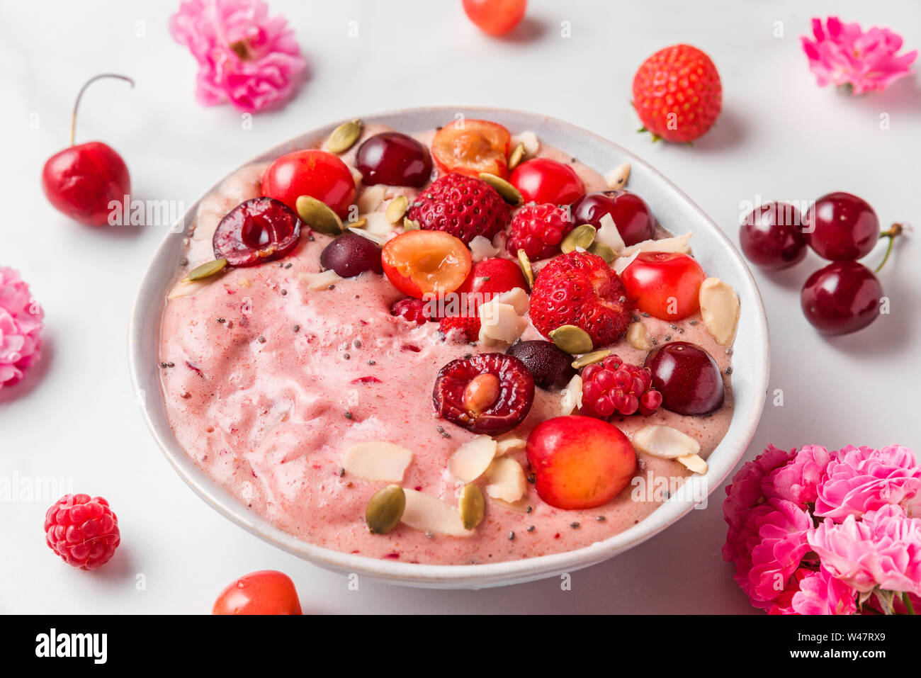 pink smoothie bowl or nice cream with fresh berries, rose flowers, nuts and seeds on white marble table. healthy breakfast. close up Stock Photo