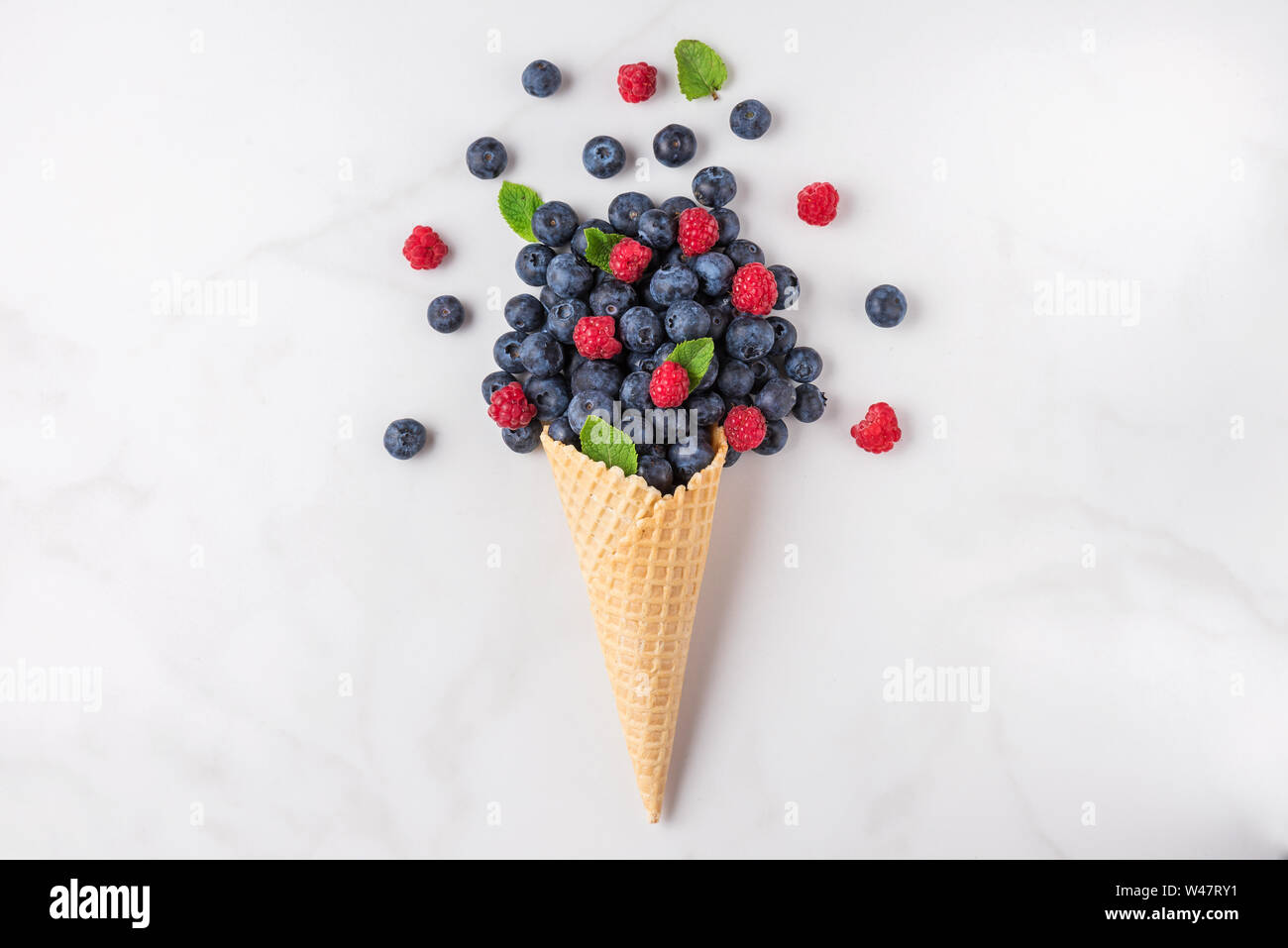 wild berries blueberry and raspberry with mint in waffle ice cream cone on white marble background. summer food concept. top view. flat lay Stock Photo