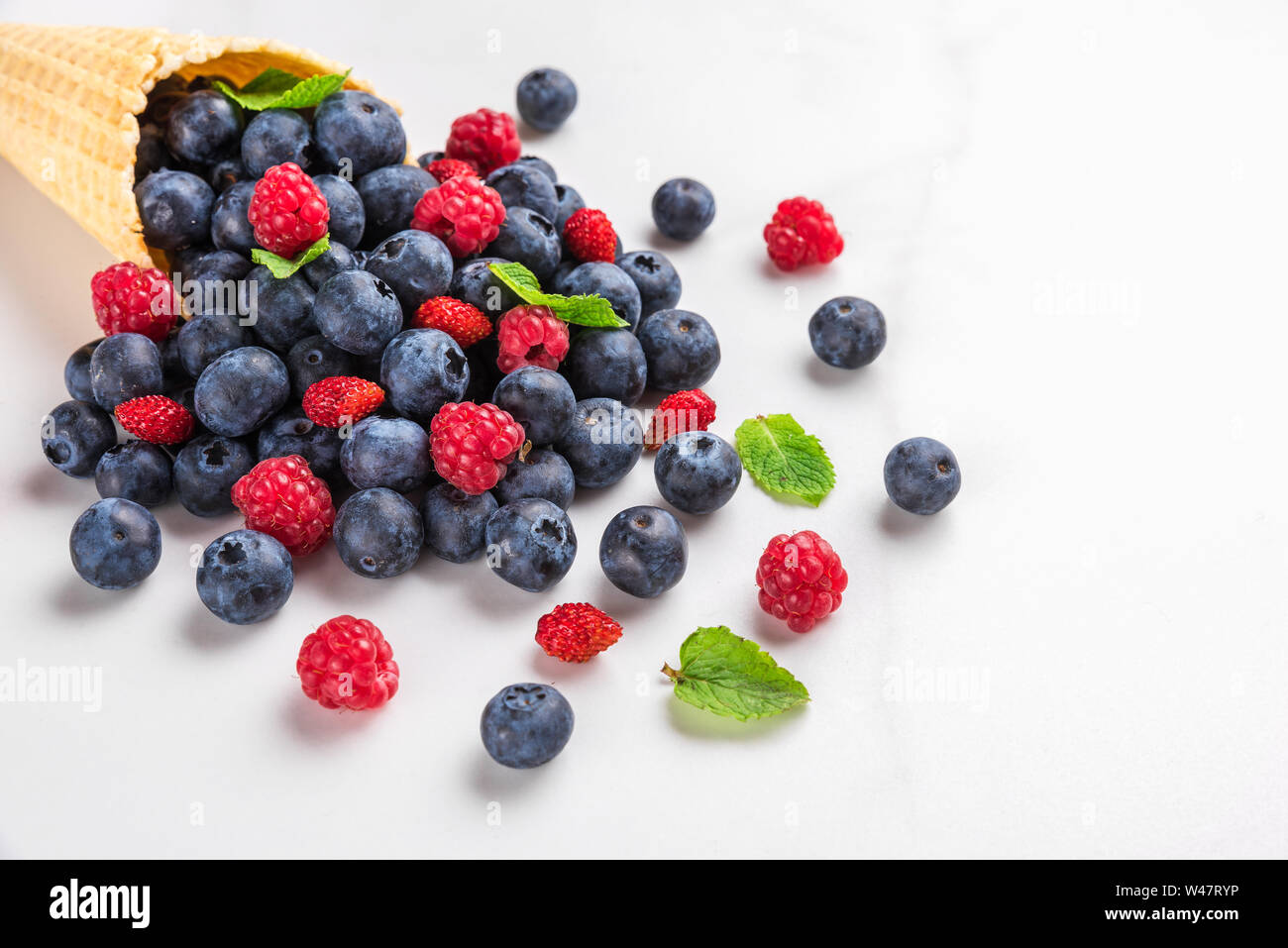 wild berries blueberry, strawberry and raspberry with mint in waffle ice cream cone on white marble background. summer food concept. close up Stock Photo