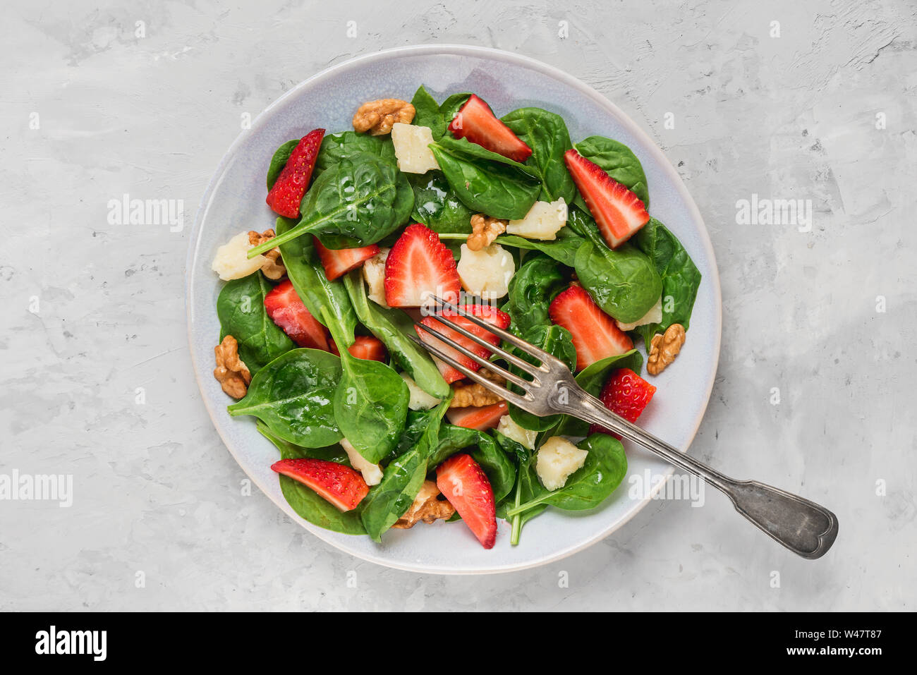 bowl of summer spinach salad with strawberry, parmesan cheese and walnuts with a fork on concrete background. healthy diet food. top view Stock Photo