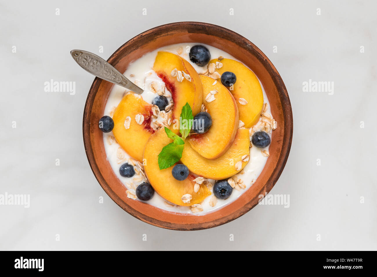 yogurt with peaches, oats, blueberries, honey and mint in a bowl with a spoon for healthy breakfast on white marble background. top view Stock Photo