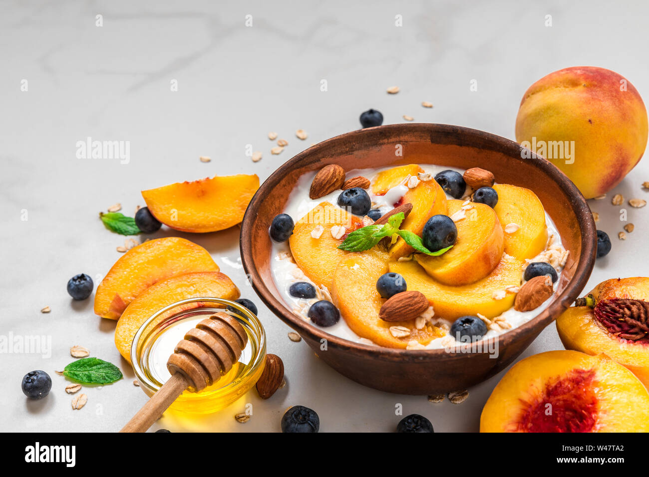 greek yogurt with peaches, oats, blueberries, honey and mint in a bowl for healthy breakfast on white marble background. close up Stock Photo