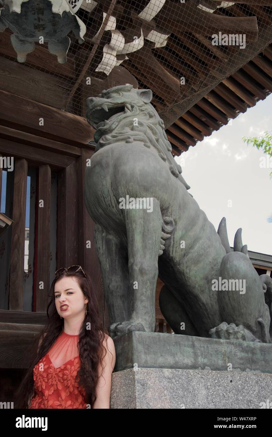A caucasian, female tourist poses with a grinning Japanese (asian) dragon (lion) statue, snarling in front of it while wearing a translucent red dress Stock Photo