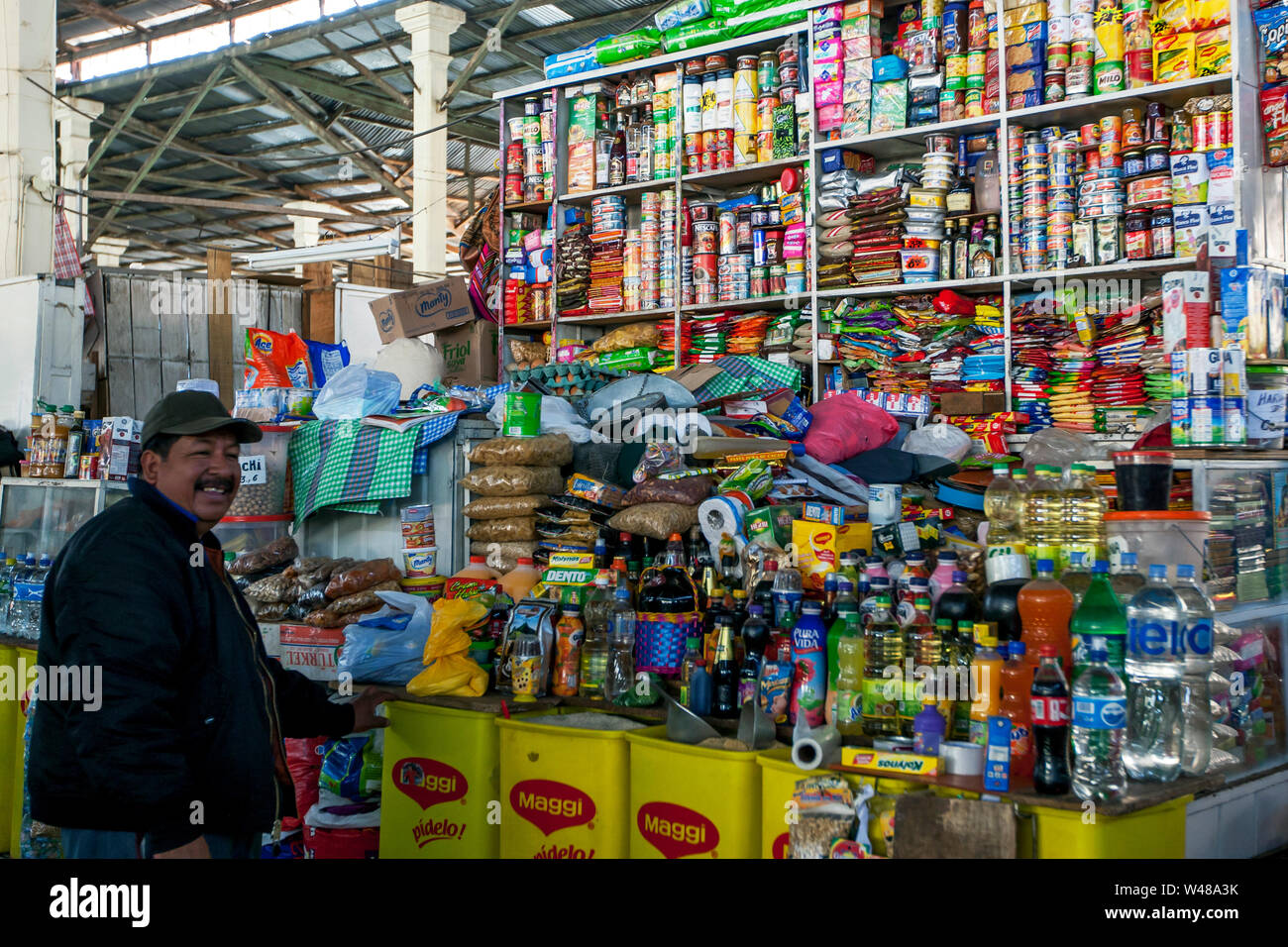 A man stands in front of his colourful stall at the undercover market in central Cusco in Peru. He sells mostly canned and manufactured produce. Stock Photo