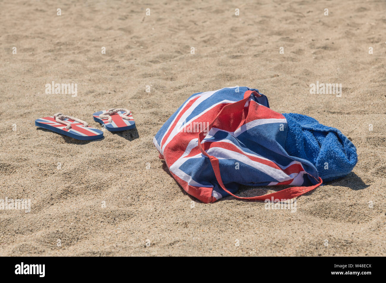 Union Jack beach bag & flip-flops on sandy beach. For 2021 UK staycation, holidays at home, staycation Cornwall, seaside holiday, flip-flop footwear. Stock Photo