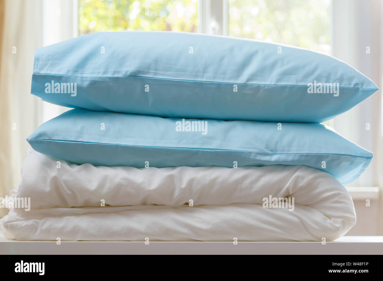 A folded duvet and pillows lie on a dresser against the background of a blurred window. Household. Stock Photo