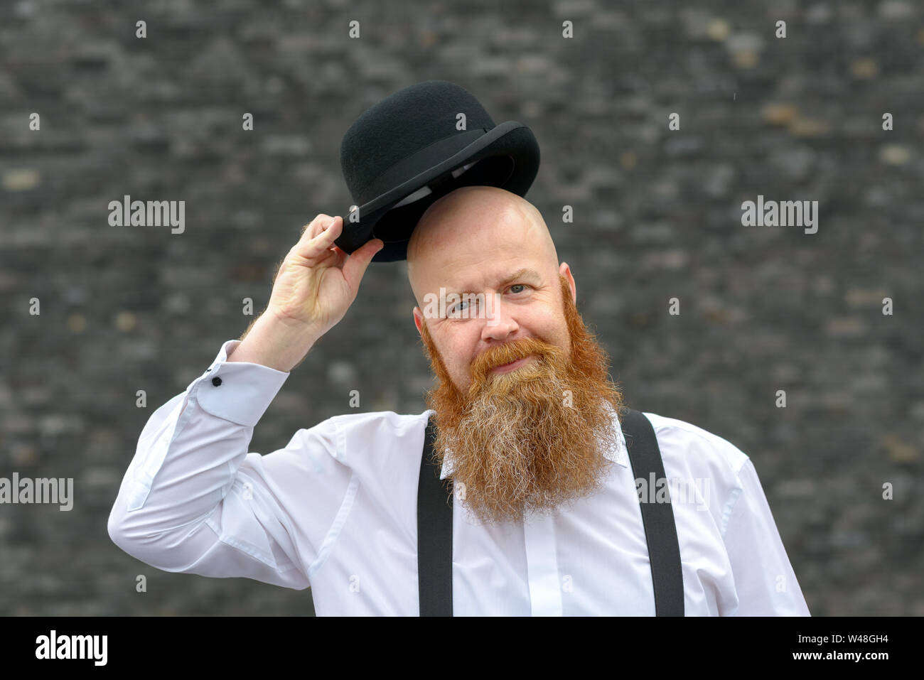 Polite bald bearded man doffing his bowler hat in greeting with a friendly welcoming smile over a grey background Stock Photo