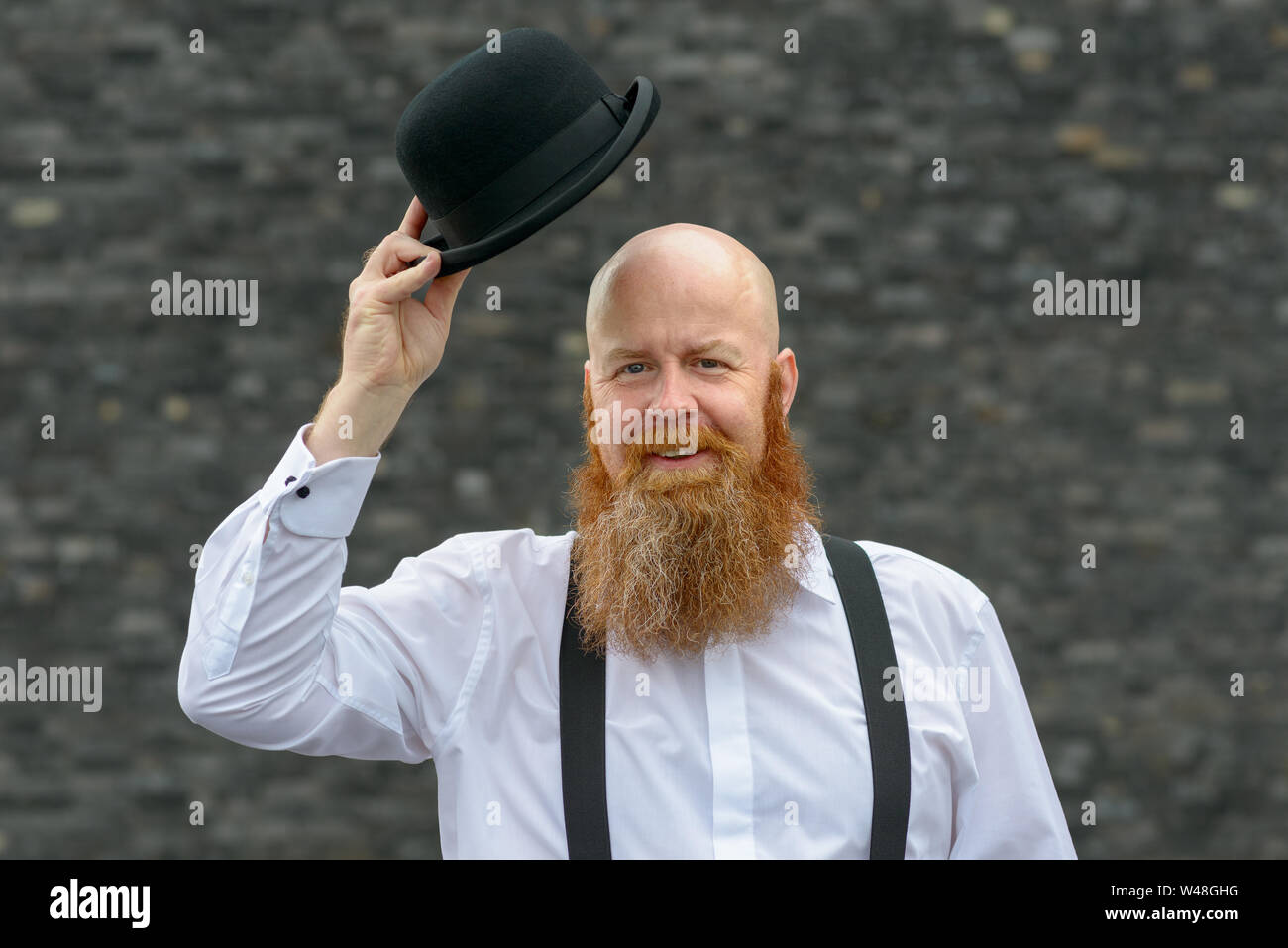 Polite bald bearded man doffing his bowler hat in greeting with a friendly welcoming smile over a grey background Stock Photo
