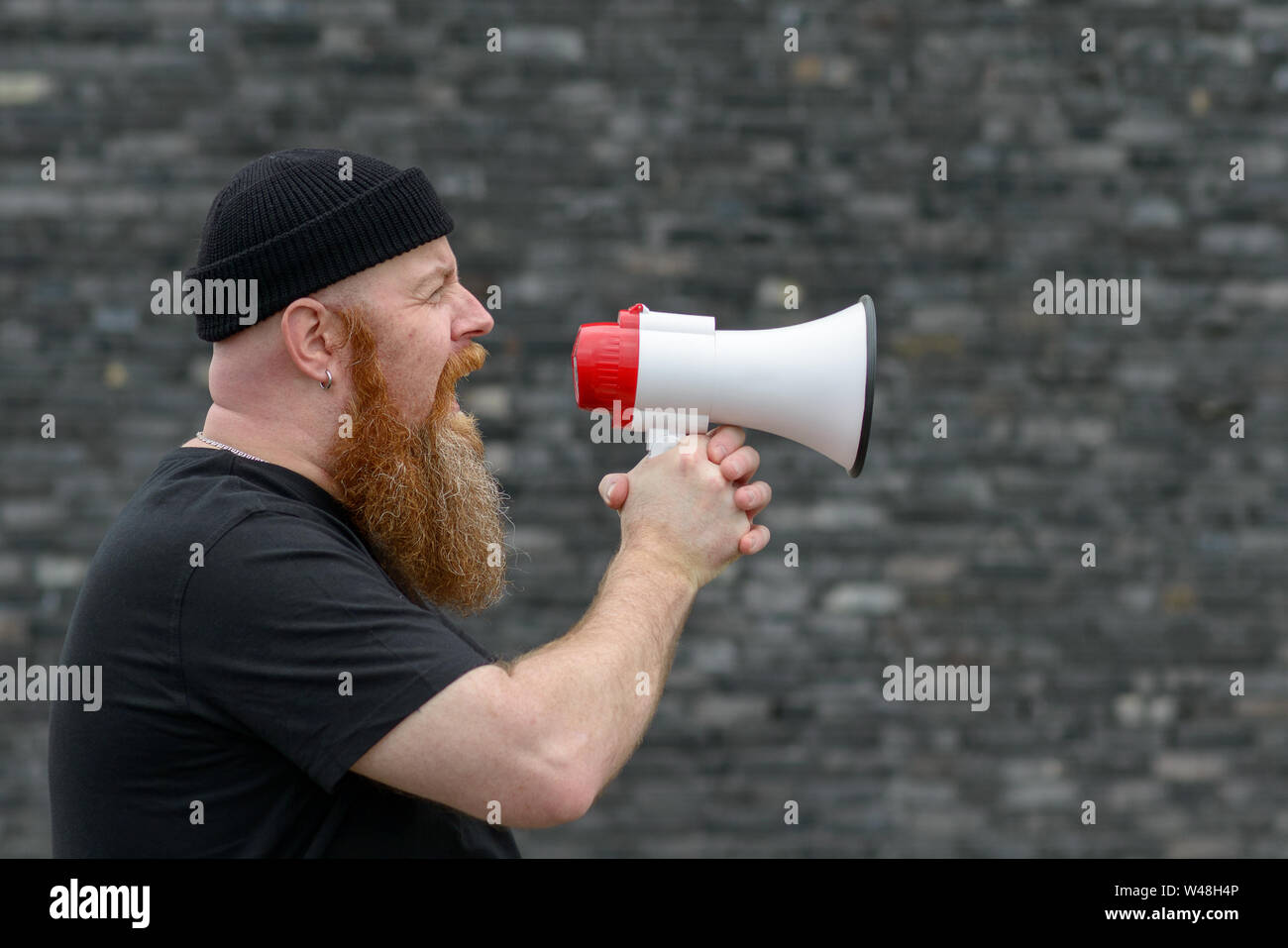 Angry bearded man in black beanie hat shouting into a megaphone airing his grievances at a strike or demonstration over a grey background Stock Photo