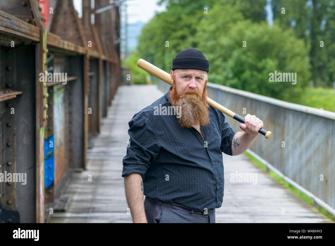 Hefty muscular bearded man with a baseball bat blocking access along a pedestrian bridge as he stares intently at the camera Stock Photo