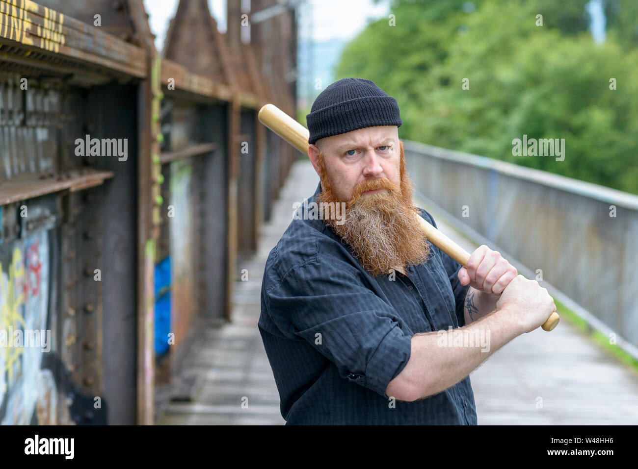 Threatening angry man with a red beard wearing a knitted beanie hat swinging a baseball bat over his shoulder while focusing his eyes on the camera Stock Photo