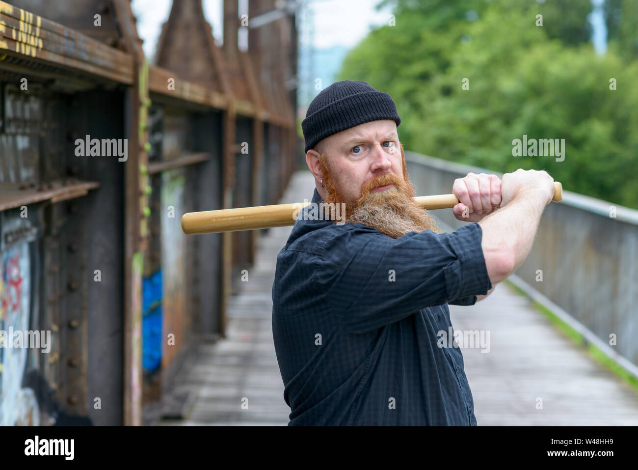 Threatening angry man with a red beard wearing a knitted beanie hat swinging a baseball bat over his shoulder while focusing his eyes on the camera Stock Photo