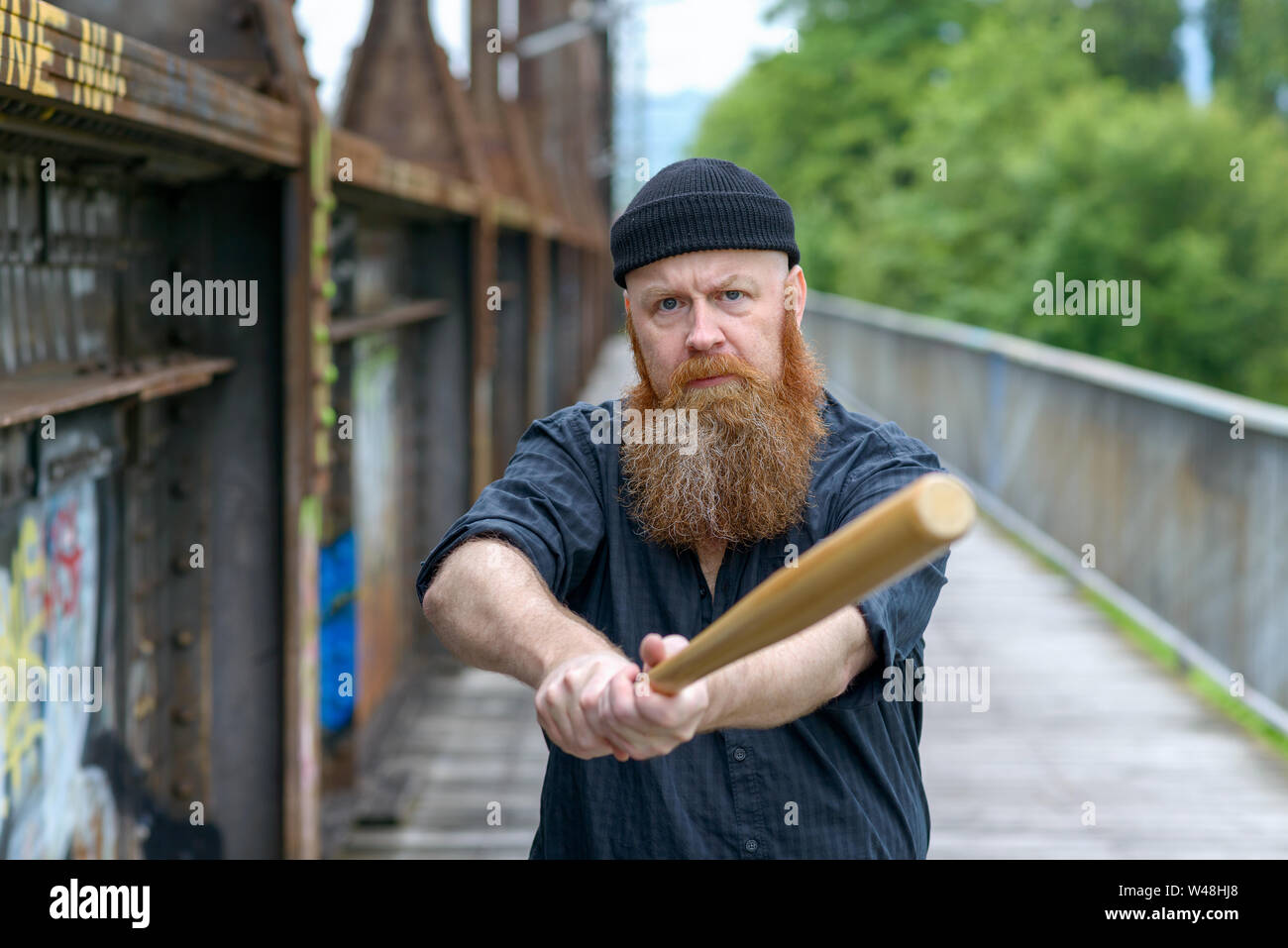 Aggressive bearded man in knitted beanie hat swinging a baseball bat at the camera with an angry expression outdoors on a bridge Stock Photo