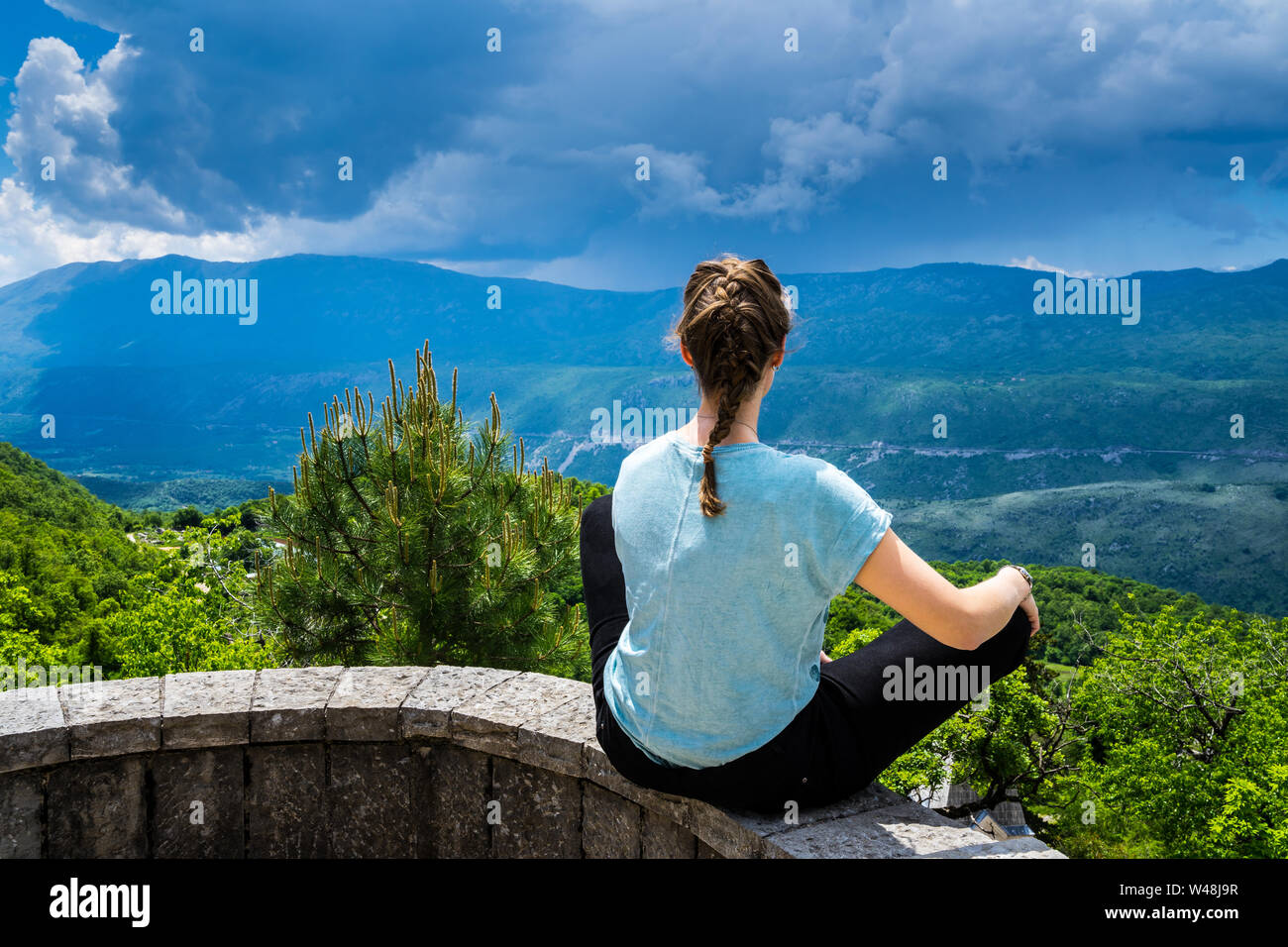 Montenegro, Beautiful young girl sitting in tailor seat on a stone wall enjoying wide view over green nature landscape bjelopavlici plain lowlands val Stock Photo