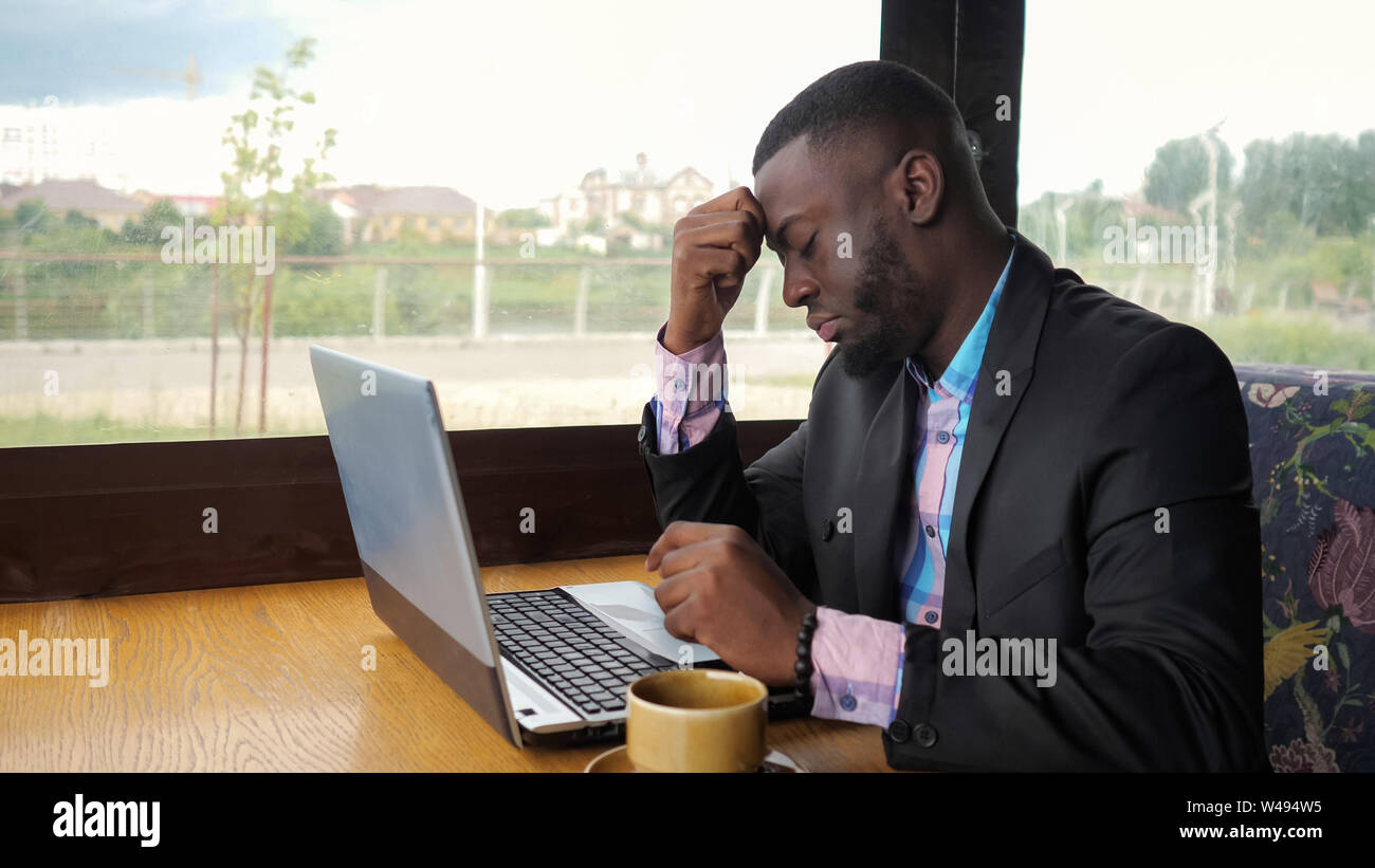 Afro american businessman tries to type on computer sitting in cafe. Black man works on laptop but it's broken and he is nervous and knocking on the keys. He has a computer problems. Battery is low. Stock Photo