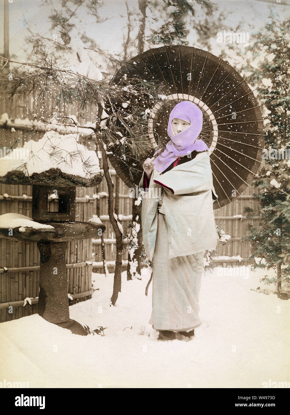 [ 1890s Japan - Japanese Woman with Parasol in the Snow ] —   A woman in a thick winter kimono has an okosozukin (御高祖頭巾) wrapped around her head to protect herself against the winter cold. She is holding a paper parasol.  19th century vintage albumen photograph. Stock Photo