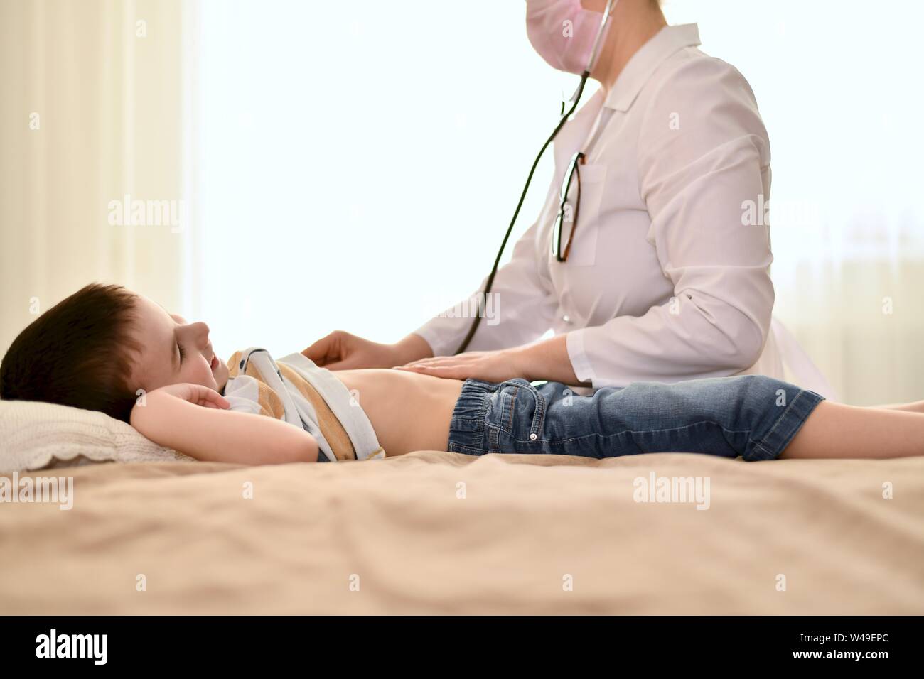 A doctor listens with a stethoscope to the heartbeat and breath of a smiling boy. The baby is at home on the bed, the doctor is sitting nearby. Stock Photo