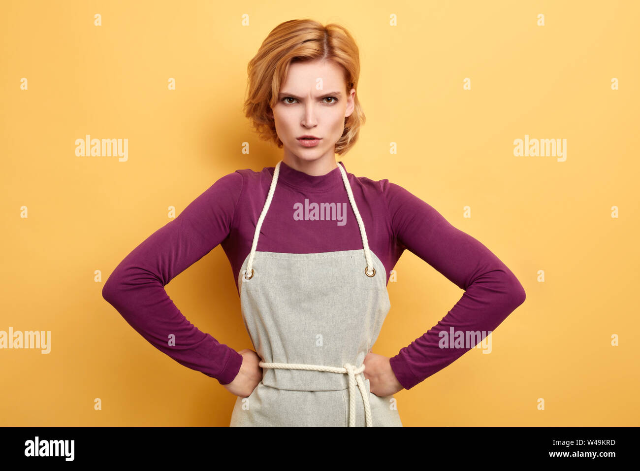 frustrated frowning housewife with hands on her hips wearing kitchen apron posing on yellow background. negative emotion and feeling. frustration and Stock Photo