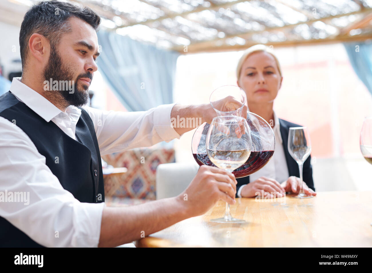 Young bearded male sommelier or bartender pouring red cabernet into wineglass Stock Photo